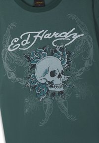 Grünes T-Shirt mit dem Text "Ed Hardy", zentraler Totenkopf umgeben von blauen Rosen sowie schwachen Flügel- und Blumendesigns im Hintergrund.