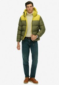 Puchowa kurtka w kolorze oliwkowym i jasnym żółtym, z kapturem, zapinana na zamek, z regulowanymi mankietami. Założona na kremowy sweter i niebieskie dżinsy.