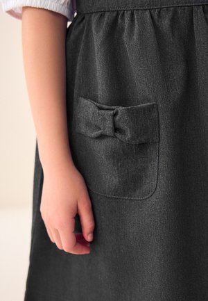 Mano di bambino appoggiata accanto a una gonna grigio scuro con una tasca frontale decorata da un fiocco di stoffa.