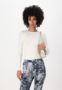 Lange ermers tilpasset kremfarget topp med "GUESS JEANS"-logo, kombinert med marineblå mønstrede bukser med blomstermotiv og teksturerte design.