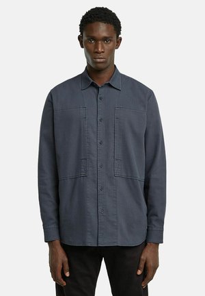 Chemise bleu foncé à manches longues avec un devant à boutons, deux poches à rabat et une coupe décontractée. Tissu lisse avec une texture subtile.
