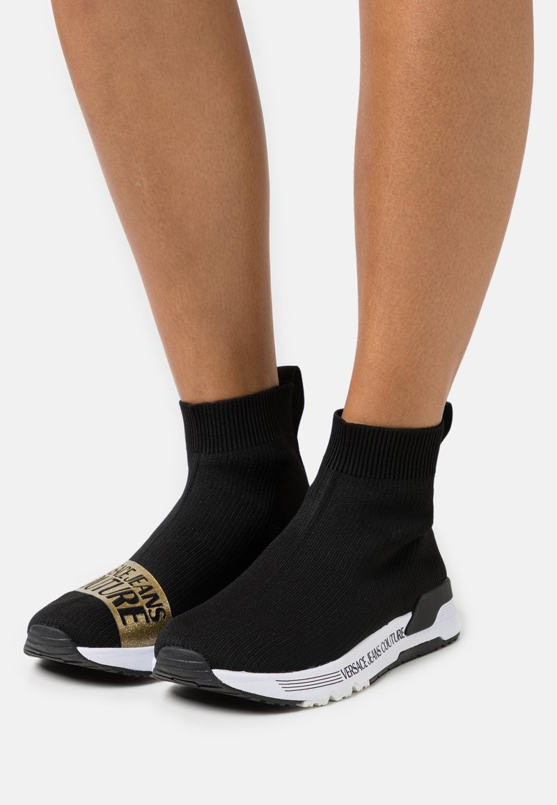 Bottines en maille noire avec un design style chaussette, dotées d'un logo doré et de semelles blanches avec des détails noirs.
