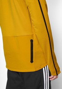 Giacca sportiva gialla con texture a coste, tasca con zip nera e striscia nera a contrasto lungo la manica, abbinata a pantaloni neri.