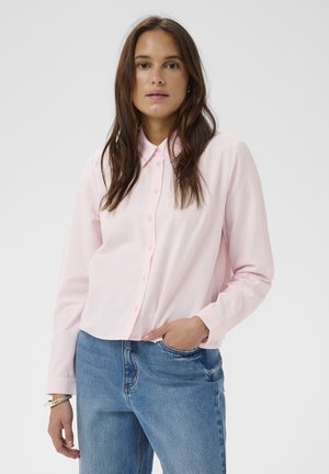 Woman with long brown hair wearing a light pink button-up shirt and blue jeans, standing with one hand in pocket against a plain background.