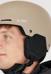 Jongere die een beige SLOPE-helmpje draagt met zwarte gewatteerde oorkappen en een verstelbare kinband, die naar links kijkt.