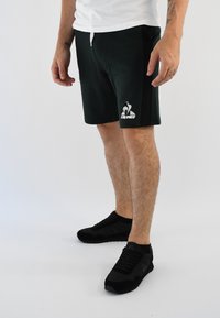 Shorts deportivos negros con cintura ajustable, con un logo blanco. Combinados con zapatillas negras, todo sobre un fondo claro.