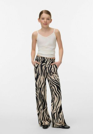Ung person stående med hænderne i lommerne iført hvidt tanktop, sort og beige zebramønstrede bukser med vide ben og sorte sko.