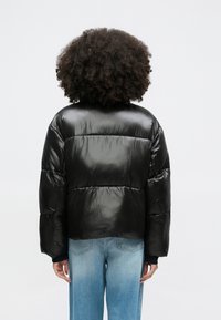 Μαύρο μπουφάν puffer με γυαλιστερή, λεία υφή, με κοντό σχέδιο και φαρδιά μανίκια. Συνδυάζεται με ανοιχτόχρωμα μπλε τζιν.