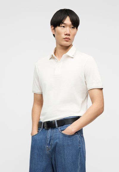 Νεαρός άντρας με μαύρα μαλλιά που φοράει λευκό κοντομάνικο πόλο μπλουζάκι, μπλε τζιν και μαύρη ζώνη, στέκεται με τα χέρια στις τσέπες.