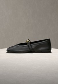Chaussure plate en cuir noir à bout pointu et à bride unique avec une petite boucle dorée, exposée sur une surface claire.