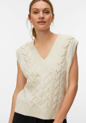 Femme portant un pull sans manches tricoté couleur crème avec un col en V et un motif de feuille texturé.