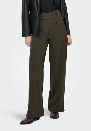Femme portant un pantalon large taille haute couleur olive foncé, un haut noir, une veste en cuir noire et des bottines à talons rouge foncé.