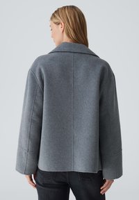 Grauer Wollmantel mit breitem Revers, überschnittenen Schultern und lockerer Passform. Verfügt über ein sauberes, minimalistisches Design mit einer leicht strukturierten Oberfläche.