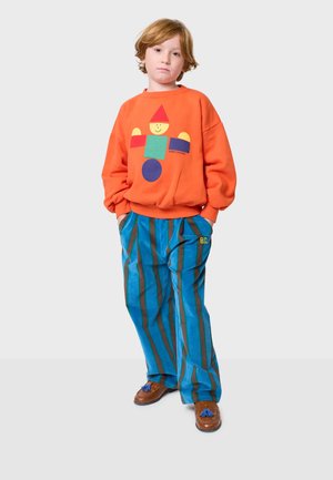 Oranges Sweatshirt mit einem bunten geometrischen Muster und blauen gestreiften Hosen aus weichem Stoff. Braune Schuhe mit blauen Quasten runden den Look ab.