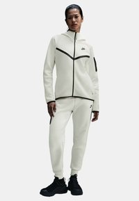 Chándal de mujer Nike en blanco roto, con cremalleras y costuras en negro que contrastan, un cuello alto y pantalones jogger ajustados con bolsillos laterales.