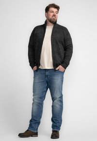 Cárdigan de carbón con cremallera y detalles acanalados, combinado con una camiseta beige clara y jeans azul desgastados. Zapatos marrones completan el atuendo.