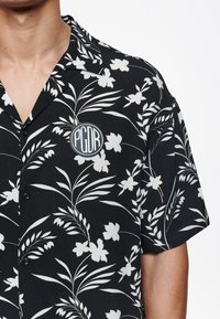 Maglietta nera a maniche corte con motivo floreale bianco, caratterizzata da una toppa circolare con il logo "PGDR" sul petto. Tessuto morbido con chiusura a bottoni.