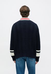 Πλεκτό μπλε ναυτικό πουλόβερ με ριμπ υφή, ριγέ λευκές και μαύρες λεπτομέρειες στα μανίκια και κόκκινο-λευκό γιακά. Συνδυάζεται με μπλε τζιν.