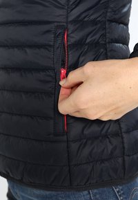 Draghand håller på att dra upp den röda dragkedjan på fickan till den svarta vadderade jackan över blå jeans.