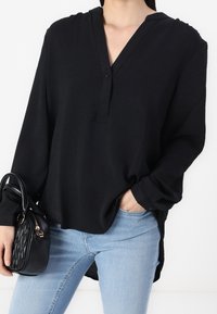 Camisa negra de manga larga con cuello en V y tapeta de botones, hecha de un tejido texturizado. Combinada con unos vaqueros azules y un pequeño bolso negro.