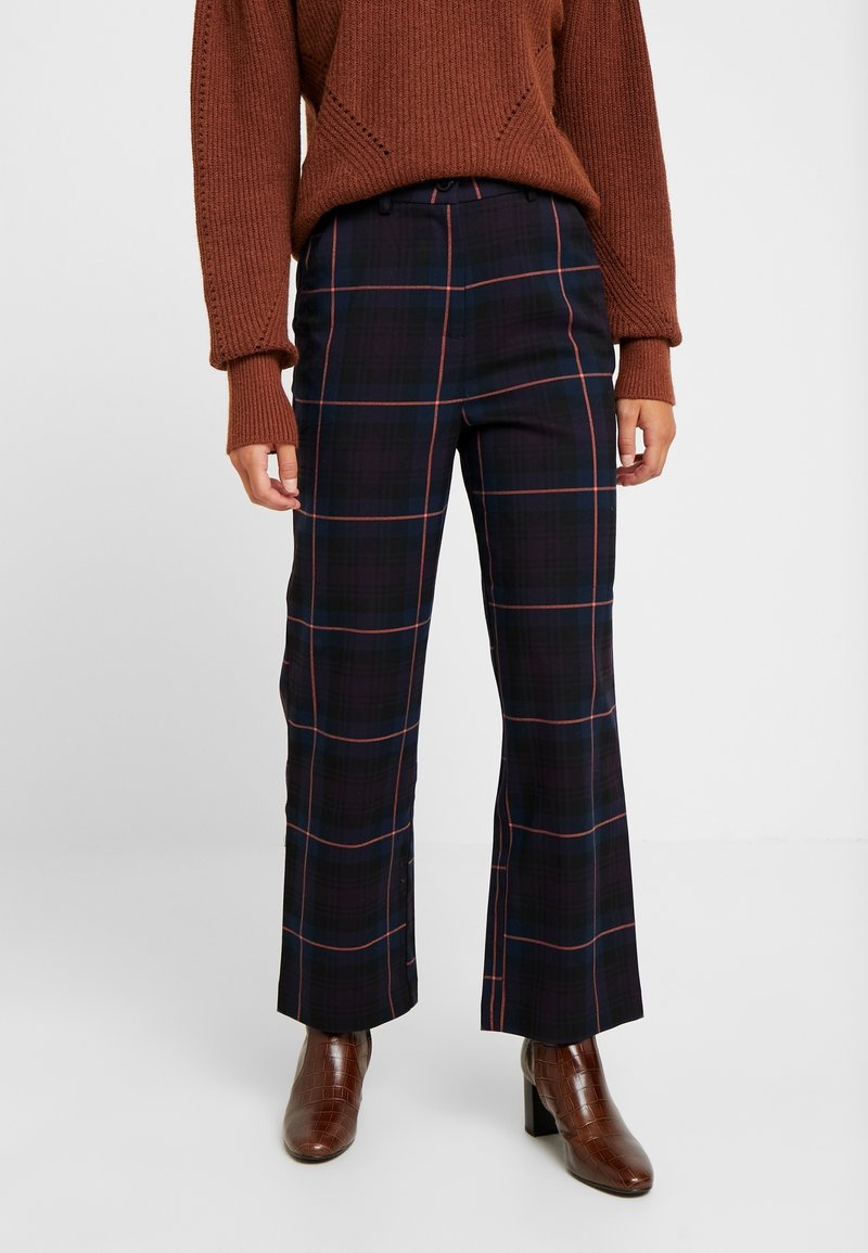 Personne portant un pull en tricot marron, un pantalon à carreaux bleu marine foncé avec des lignes rouges et bleues, et des bottines marron à talons, se tenant devant un fond uni.