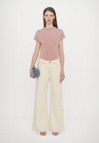 Top scurt cu mâneci scurte în nuanță de piersică, asortat cu blugi largi crem; modelul ține în mână o geantă mică albastră și poartă sandale bej. Design simplu și modern.