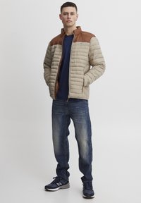 Polstret jakke i beige med brune skulderdetaljer, med lynlås lukning. Bæres med en mørkeblå skjorte og denim jeans. Sorte sneakers fuldender looket.
