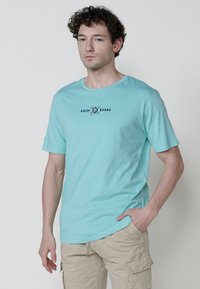 Helles blaues T-Shirt mit kurzen Ärmeln aus weichem Stoff, das ein schwarzes Grafikdesign und den Text "KRASHI BRAND" auf der Vorderseite aufweist. Klassische Passform.