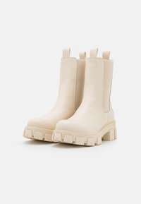 Bottines beiges en cuir lisse, dotées de panneaux latéraux élastiques, d'une semelle épaisse en caoutchouc texturé avec des rainures, et de languettes de tirage.