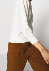 Pull blanc en maille avec un motif texturé, col rond et manches trois-quarts, associé à un pantalon marron sur mesure.