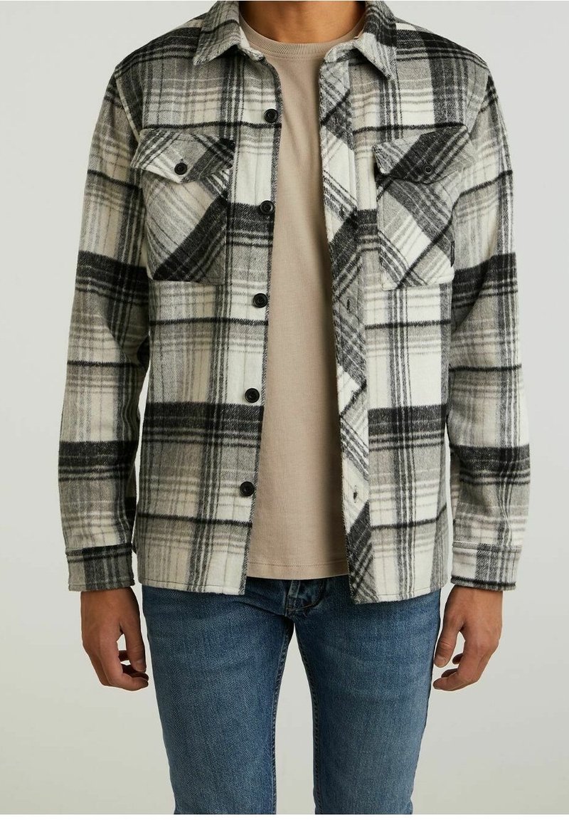 Ruutupaita tummissa väreissä, mustassa, valkoisessa ja harmaassa, joissa on kaksi rintataskua, napillinen etusuljenta ja pitkät hihat. Päälle puettuna beigellä paidalla.