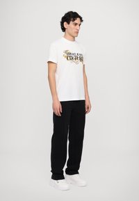 Balta medvilninė marškinė su grafiniu spaudiniu "VERSACE JEANS COUTURE" juoda ir geltona spalvomis, derinami su juodomis kelnėmis ir baltomis sportbačiais. Minimalistinio dizaino.