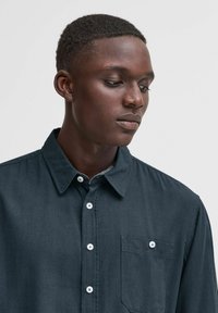 Camicia button-up blu scuro in un tessuto morbido, con colletto a punta, taschino frontale e bottoni bianchi; texture liscia.