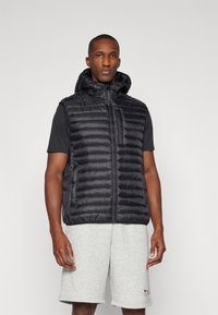 Črn puhovka z kapuco, z žakardnim dizajnom, z zadrgo in stranskimi žepi, nosi se preko sivega majice in kratkih hlač.