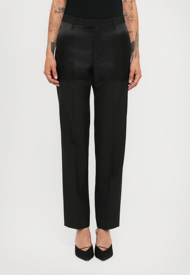 Pantalons droits noirs ajustés avec des détails en patch sur les cuisses supérieures, portés avec des chaussures noires à bout pointu et des bras tatoués visibles.