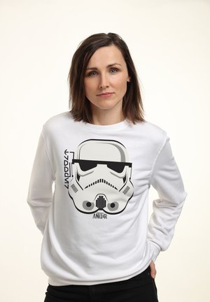 Kobieta o brązowych włosach do ramion nosząca białą bluzę z czarną grafiką hełmu szturmowca i słowem "ANDOR."