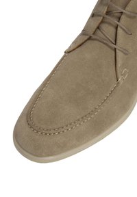 Zapato de ante beige claro con punta redondeada, detalle de costura en capas a lo largo del borde y suela de goma de color claro. Cordones visibles en la parte superior.
