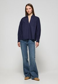 Camicia a maniche lunghe navy con colletto, dotata di tasca frontale e maniche plissettate. Abbinata a jeans a zampa d'elefante azzurri e scarpe beige.