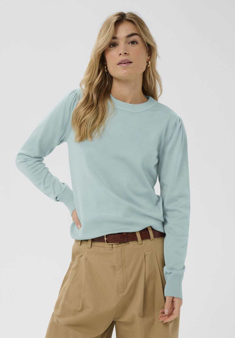 Femme aux longs cheveux blonds portant un pull à manches longues bleu clair, une ceinture marron et un pantalon taille haute beige sur un fond uni.