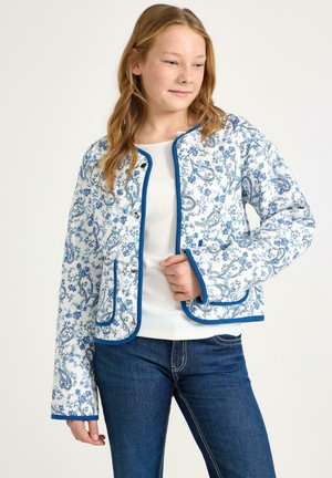 Ung kvinde iført en hvid jakke med blå blomsterprint og blå kant, kombineret med en hvid top og blå jeans, stående mod en ensfarvet baggrund.