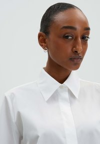 White collared shirt with a button-down front, smooth fabric texture. The model wears small, circular earrings with clear accents.