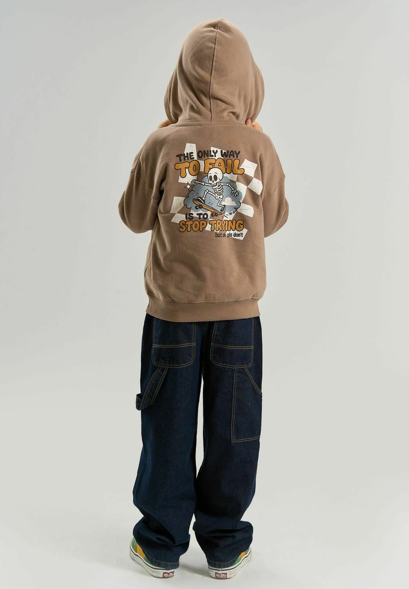 Enfant portant un sweat à capuche beige avec un squelette en skateboard et le texte « La seule façon d'échouer est d'arrêter d'essayer » dans le dos, pantalon large foncé et baskets.