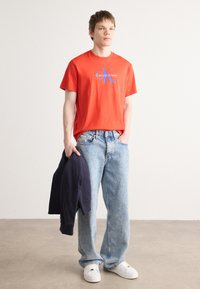 Červené bavlnené tričko s modrým logom "Calvin Klein", v kombinácii so svetlomodrými voľnými džínsami a bielymi teniskami. Čierna bunda držaná v ľavej ruke.