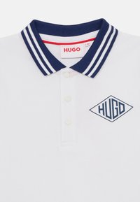 Bela polo majica z ovratnikom v mornarsko modrih črtah, ki ima modri logotip "HUGO" v obliki diamanta in tri gumbe. Teksturni material.