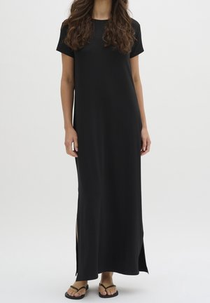 Kobieta ubrana w prostą czarną maxi sukienkę z krótkim rękawem i rozcięciami po bokach, w czarnych japonkach, stojąca na białym tle.