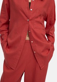 Osoba w luźnej, czerwonej koszuli zapinanej na guziki z kieszonką na przodzie oraz pasujących czerwonych spodniach, z rękami w kieszeniach, widoczny środkowy odcinek ciała.