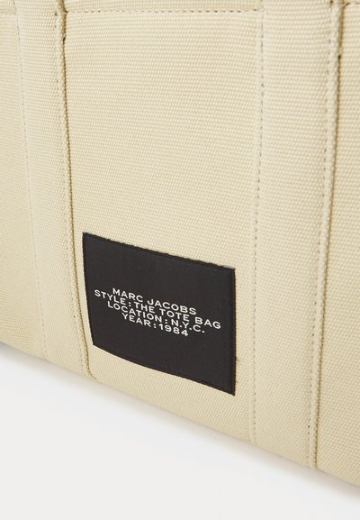 Beige canvas shoppingväska med en strukturerad yta, prydd med en svart etikett med vit text som specificerar varumärke och stilinformation.