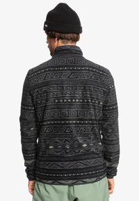Fleece-pullover i svart med gröna geometriska mönster, med krage, långa ärmar och ett strukturerat tyg för värme och komfort.