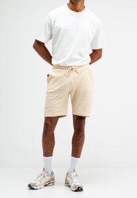 Beige strukturierte Shorts mit Kordelzugbund, kombiniert mit einem schlichten weißen T-Shirt und gemusterten Sportschuhen, getragen über weißen Socken.