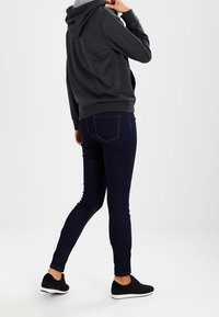 Donkergrijze hoodie met een capuchon, gecombineerd met donkerblauwe skinny jeans en zwarte sneakers, gezien vanuit de achterkant tegen een lichtgrijze achtergrond.